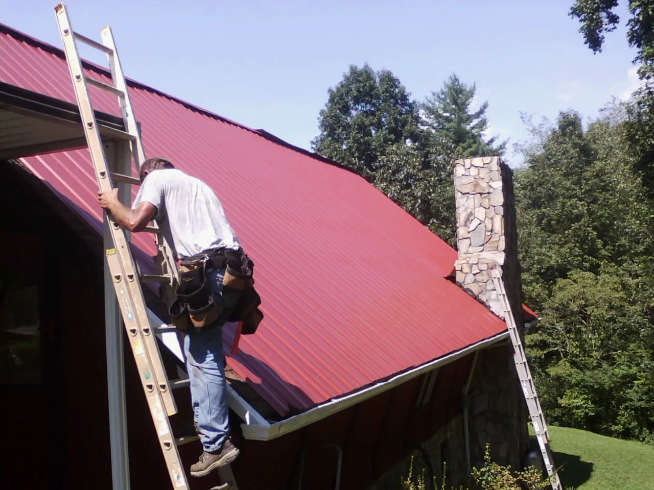 Roofer on a ladder beside a red metal roof and stone chimney — Soffit & Fascia Repair by Roofing Brothers in Pahrump
